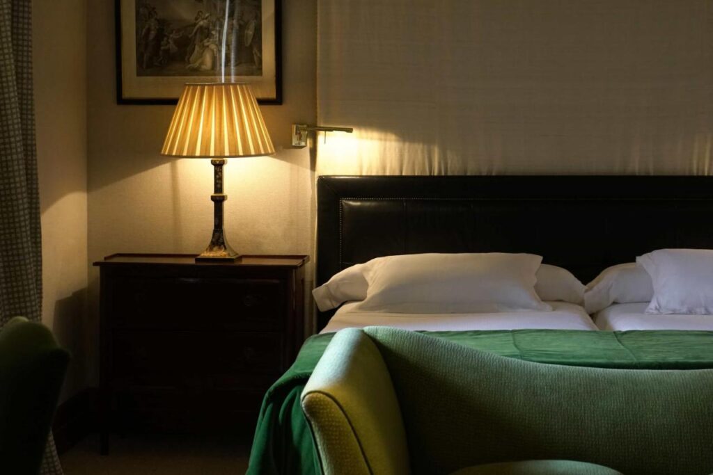 25 Weekend Trips from Madrid Max. 2h Away 54 A cozy hotel room perfect for weekend trips from Madrid, featuring a bed topped with white pillows, a green blanket, and a dark headboard. A lamp with a pleated shade softly lights the space, while a framed picture hangs above.