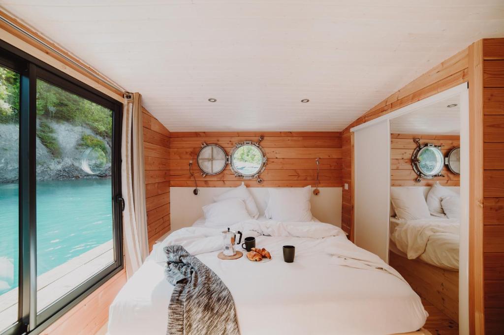 11 Unique Places to Stay in Provence 19 A cozy bedroom with wood-paneled walls, a large bed with white bedding, porthole windows, and a mirrored closet—one of the unique places to stay in Provence. A sliding glass door opens to stunning views of turquoise water outside.