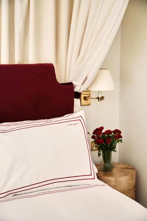 15 Best Boutique Hotels in London, UK 6 A cozy bedroom corner inspired by the best boutique hotels London, with a white pillow trimmed in red on a bed with a burgundy headboard, cream curtains, a wooden nightstand holding red flowers, and a wall-mounted lamp.