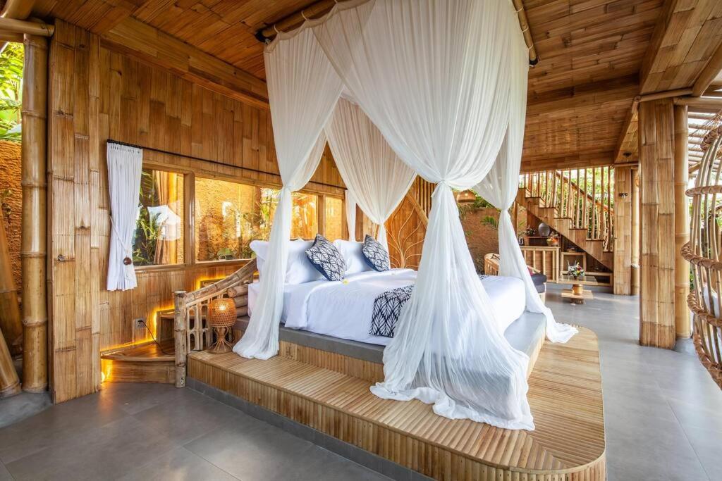 A cozy bedroom in one of the best family villas in Bali, with bamboo walls and furniture, features a bed with white linens and a flowing canopy. A staircase leads upstairs, and natural light brightens the airy, eco-friendly space.