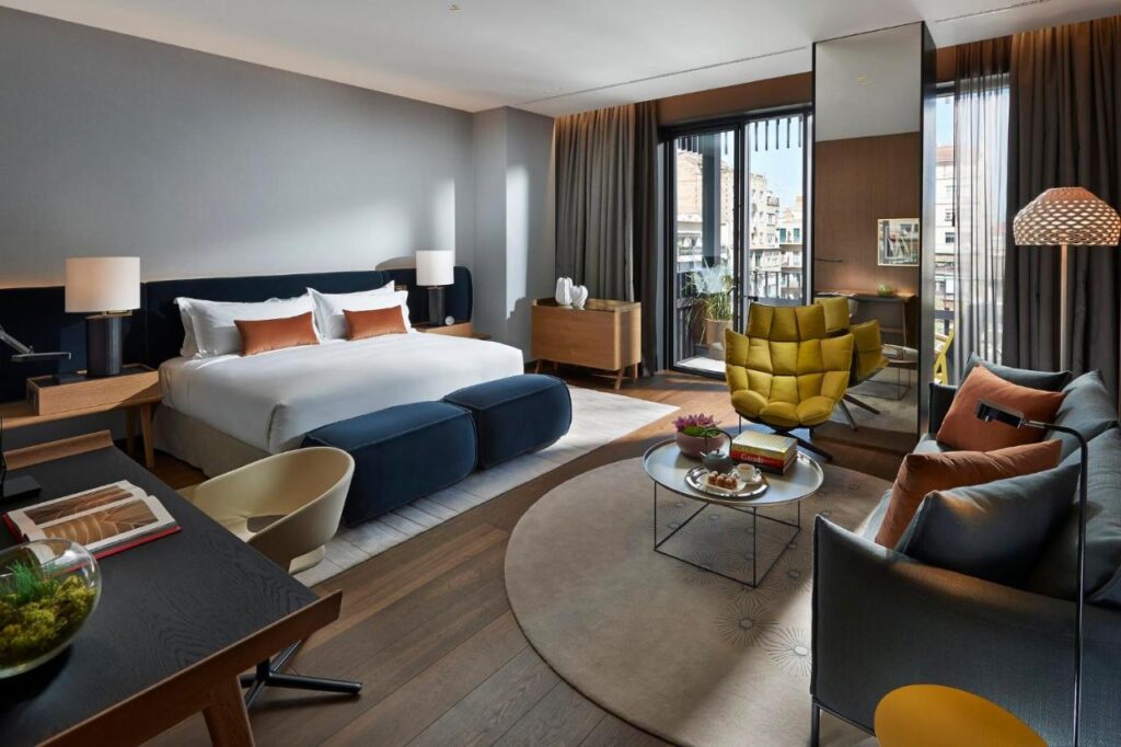 Top Barcelona Hotels with Rooftop Pool 18 Modern hotel room with a king-size bed, desk, yellow chair, sofa, round coffee table with snacks, large windows overlooking the city, and neutral decor with orange and gold accents—ideal when exploring Barcelona hotels with rooftop pool amenities.