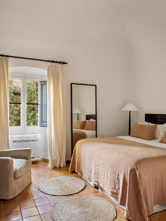 A cozy bedroom inspired by boutique hotels in Costa Brava, featuring a double bed with beige linens, a large mirror, gray armchair, woven rugs, terracotta tiles, and a window with white curtains letting in natural light.