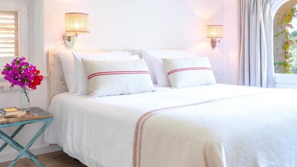 A bright, cozy bedroom reminiscent of boutique hotels in Costa Brava, with a neatly made bed featuring white linens and red-striped pillows, wall-mounted lamps, a blue side table with pink flowers, and a window with light curtains.