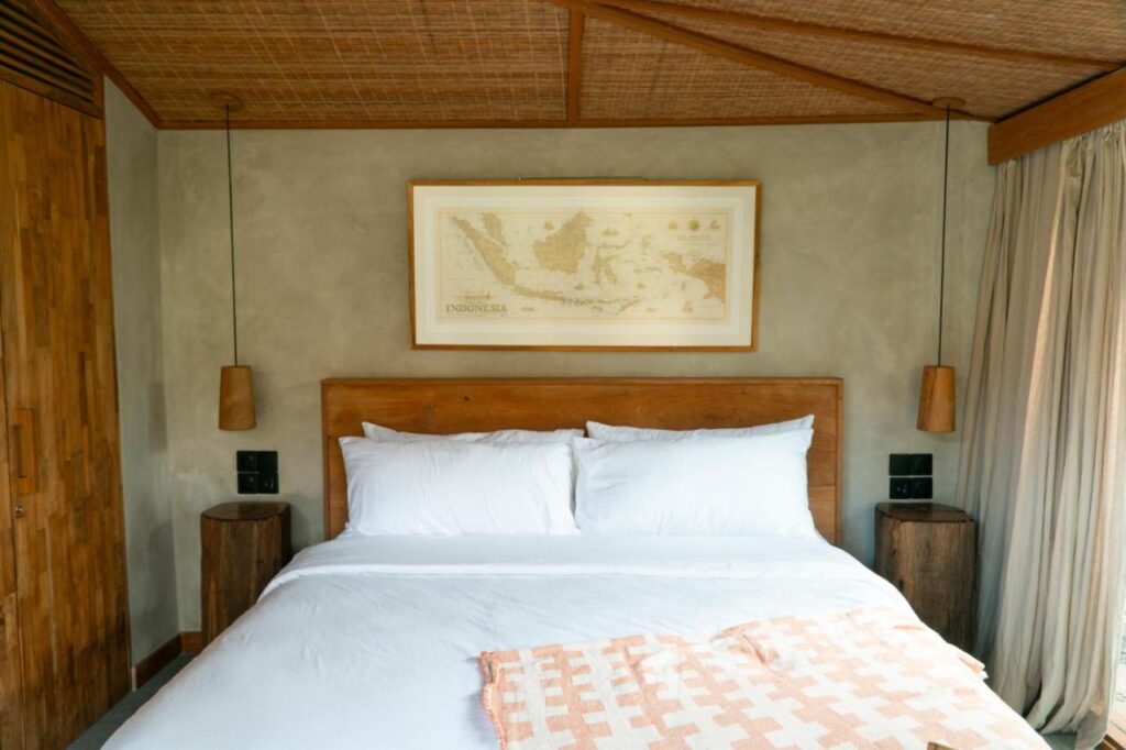 A cozy bedroom in one of the best family villas in Bali, featuring a large wooden bed with white bedding, round nightstands, hanging pendant lights, a woven ceiling, and a framed map of Indonesia above the bed. Light filters through beige curtains.