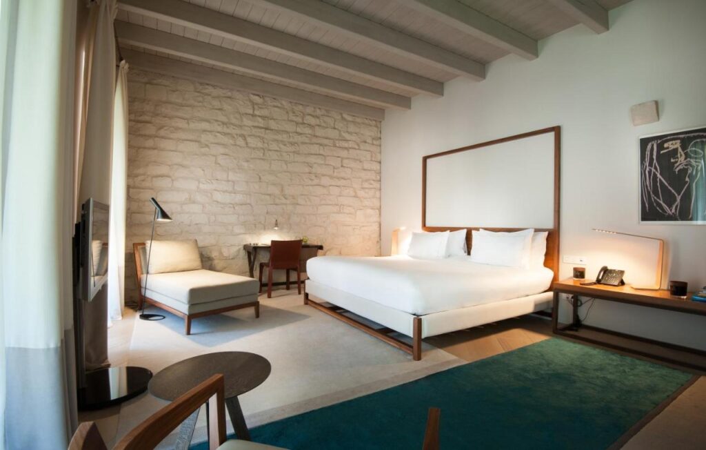 Top Barcelona Hotels with Rooftop Pool 14 A modern hotel room with a large bed, desk, lounge chair by the window, exposed stone wall, and wooden beams—perfect for relaxing after exploring Barcelona Hotels with Rooftop Pool. Soft lighting and neutral tones create a welcoming atmosphere.