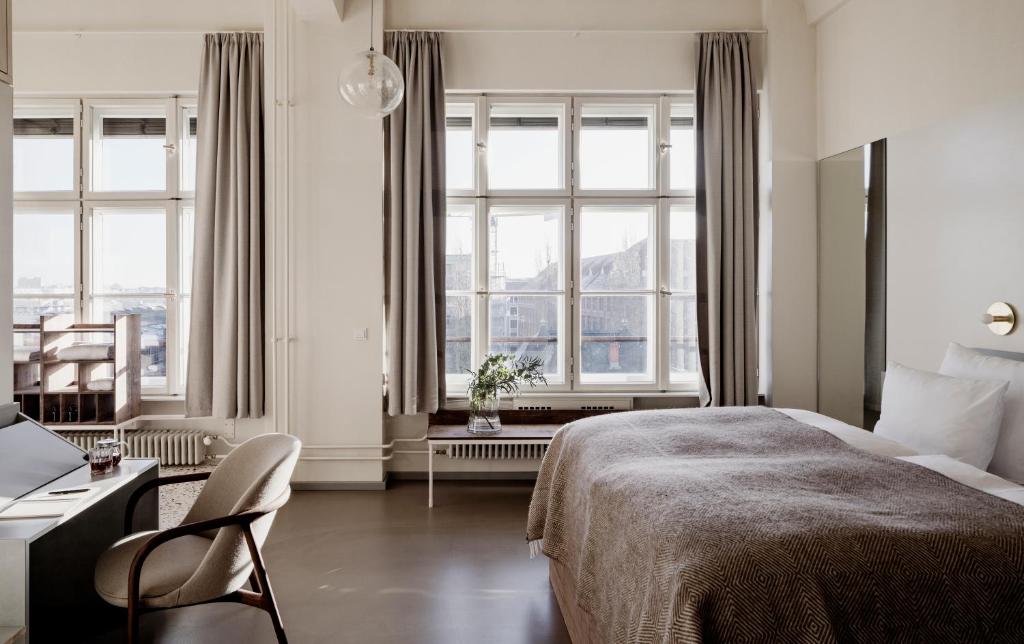 Boutique Hotels in Berlin Kreuzberg 11 A bright, minimalist bedroom inspired by boutique hotels in Berlin Kreuzberg, featuring large windows, light gray curtains, a neatly made bed with a brown blanket, a desk with a chair, and a plant on the windowsill filled with natural light.