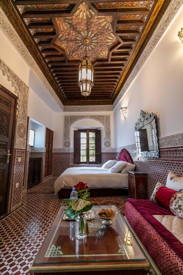 7 UNIQUE Riads in Fez You Won't Forget 29 A luxurious Moroccan bedroom inspired by riads in Fez features intricate tile flooring, ornate woodwork, a carved ceiling, a grand chandelier, a plush bed, red velvet sofa, and a decorated table with flowers and snacks as sunlight streams through the window.