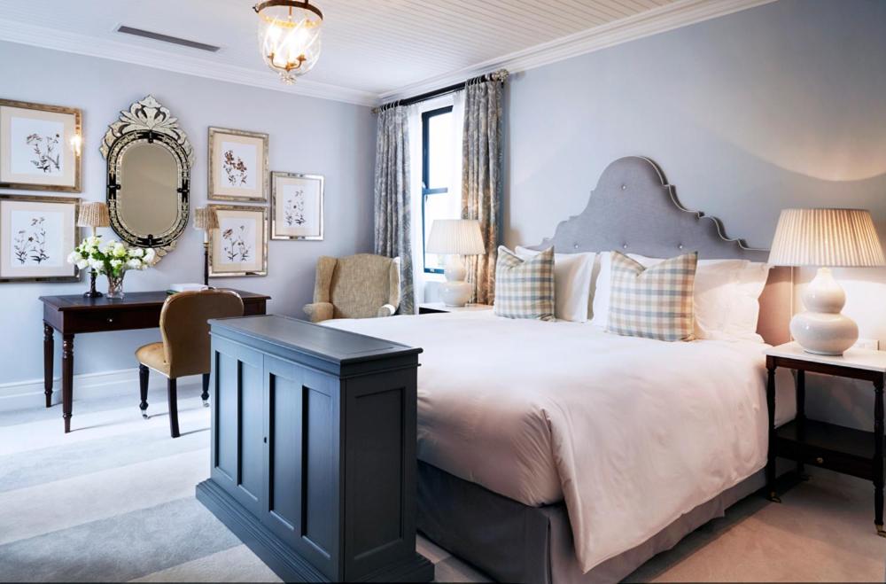 Unique Places to Stay in Cape Town 9 Elegant bedroom with a large bed, gray upholstered headboard, checkered pillows, and framed art on light blue walls—one of the unique places to stay in Cape Town, complete with a writing desk, armchair, bedside lamps, and window with gray curtains.