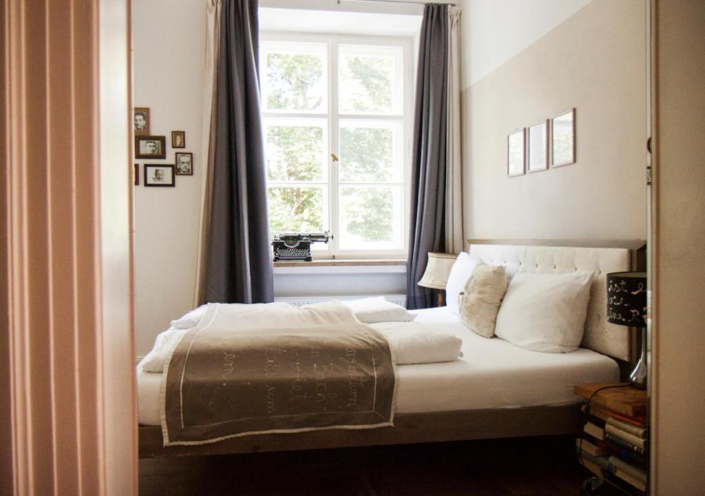 FAIRYTALE Castle Hotels in Bavaria 23 A cozy bedroom with a neatly made bed, white bedding, and gray throw blanket near a large window with dark curtains evokes the charm of castle hotels in Bavaria. A vintage typewriter sits on the windowsill, and framed pictures hang on the wall.