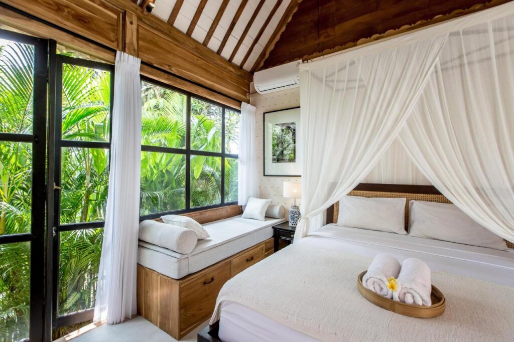 14 UNIQUE Boutique Hotels in Canggu, Bali 42 A bright, airy bedroom with a canopy bed, white linens, and rolled towels on a tray. Large windows reveal lush green plants outside—evoking the relaxing vibe of boutique hotels in Canggu. A window seat and framed photo add to the cozy, tropical feel.