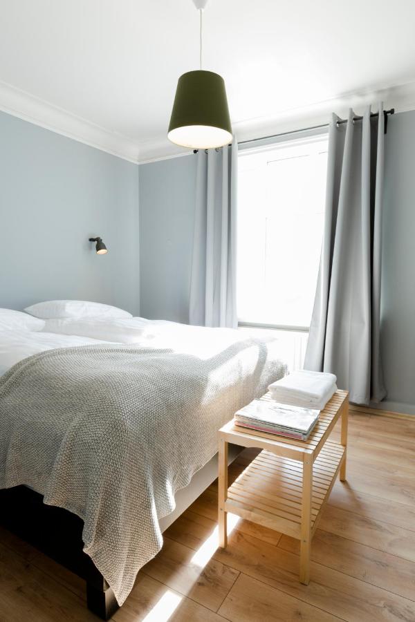 Where to Stay in Reykjavík: Top Guide 18 A bright, minimalist bedroom with light blue walls, a large bed with white bedding and a gray blanket—perfect for travelers searching where to stay in Reykjavik. Wooden floors, gray curtains, a small bench, and a green ceiling lamp complete the serene space.