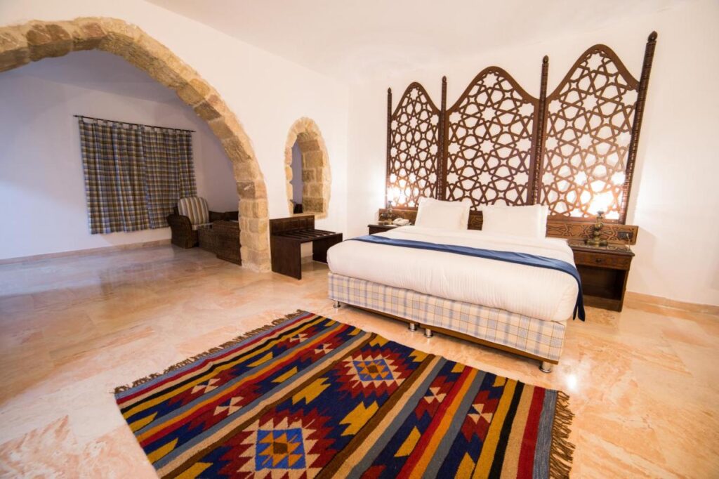 Authentic Hotels in Petra Jordan: Stay near the Ancient City 1 A spacious bedroom found in many hotels in Petra, Jordan, featuring a large bed with an intricate wooden headboard, colorful patterned rug, stone archways, wicker chairs and table in the corner, and a light polished stone floor.