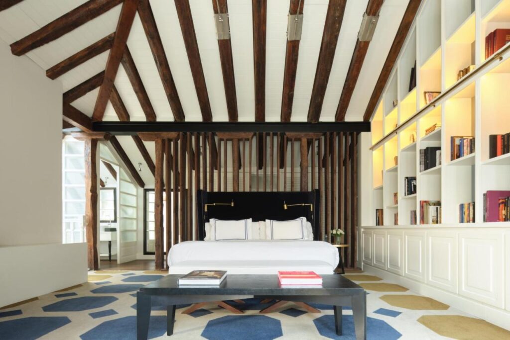 Where to Stay in Madrid: 14 Best Hotels 44 A modern bedroom, perfect for those seeking where to stay in Madrid first time, features exposed wooden beams, a large bed with white linens and black headboard, built-in bookshelves, a geometric rug, and a sleek black bench lined with books.