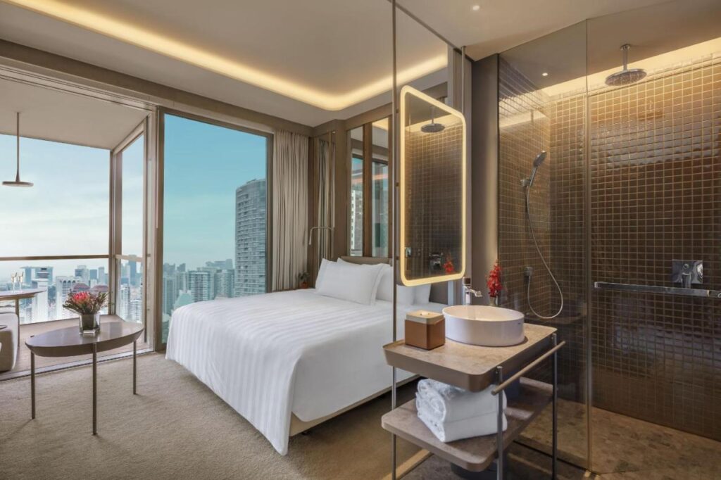 13 Most Unique Hotels in Singapore for an Incredible Holiday 42 Modern hotel room with a large bed, floor-to-ceiling windows overlooking a city skyline, and a glass-enclosed bathroom. Experience one of the unique hotels in Singapore, featuring neutral tones and soft lighting throughout.