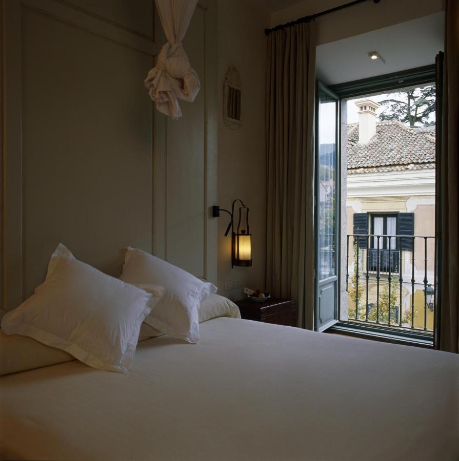 25 Weekend Trips from Madrid Max. 2h Away 90 A neatly made bed with white pillows is beside an open window, letting in natural light and offering a view reminiscent of charming stays on weekend trips from Madrid. A wall lamp and curtains frame the cozy room.