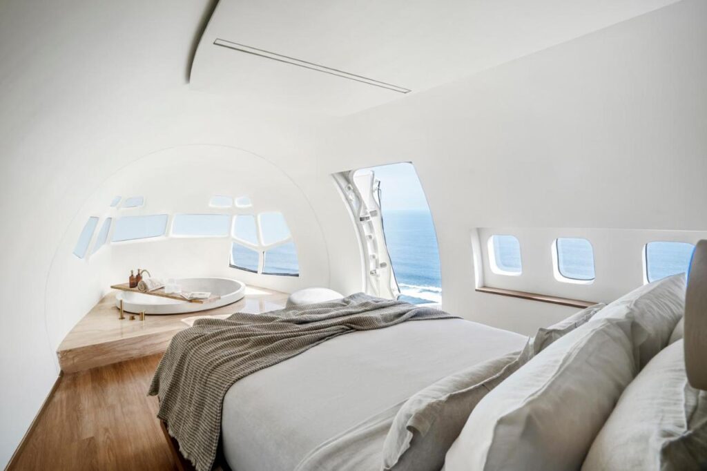 A modern bedroom with white walls and wood floors inside an airplane fuselage, featuring a cozy bed with gray bedding and large windows offering a stunning ocean view—just like the best family villas in Bali.