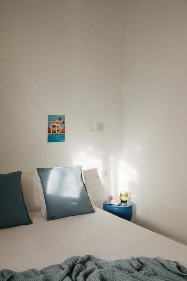11 UNIQUE Boutique Hotels in Tenerife 9 A sunlit, minimalist bedroom reminiscent of boutique hotels in Tenerife features a bed with white and blue pillows, a textured blue blanket, and a small nightstand holding a green glass. A postcard is taped to the crisp white wall.