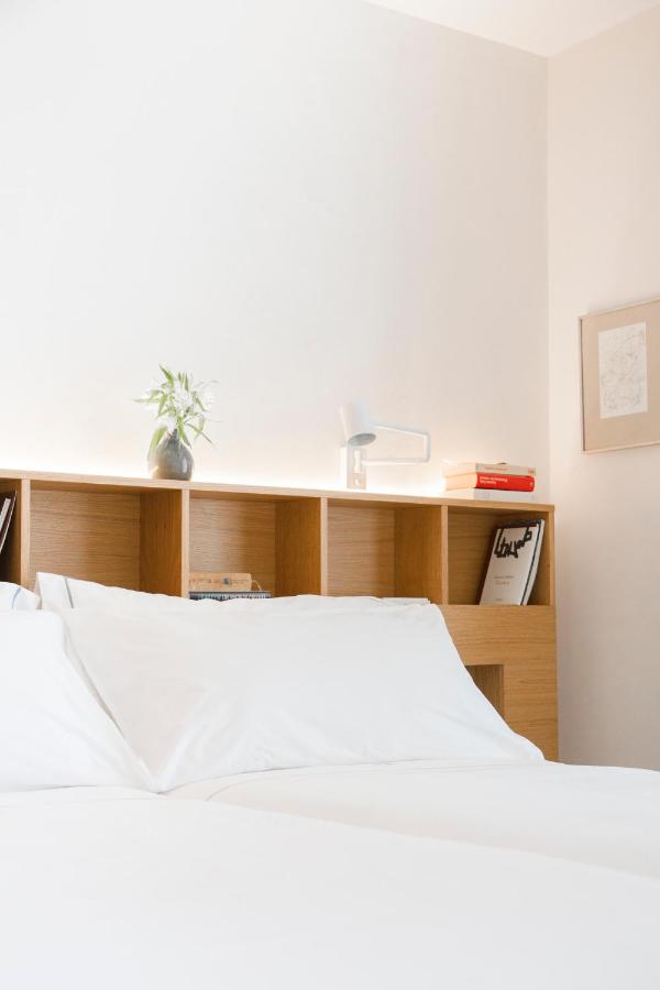 25 Weekend Trips from Madrid Max. 2h Away 64 A neatly made bed with white linens sits in front of a wooden headboard with shelves—perfect for planning weekend trips from Madrid, with books, a small green plant in a vase, a white lamp, and a framed picture on the wall.