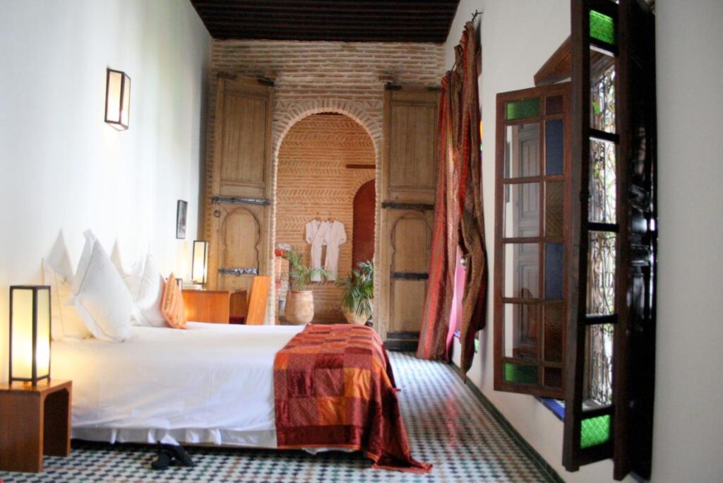 7 UNIQUE Riads in Fez You Won't Forget 24 A bright, cozy bedroom inspired by riads in Fez, featuring a large white bed, patterned tile floor, wooden furniture, open stained glass windows, and an archway leading to a rustic alcove with plants and decorative robes on display.