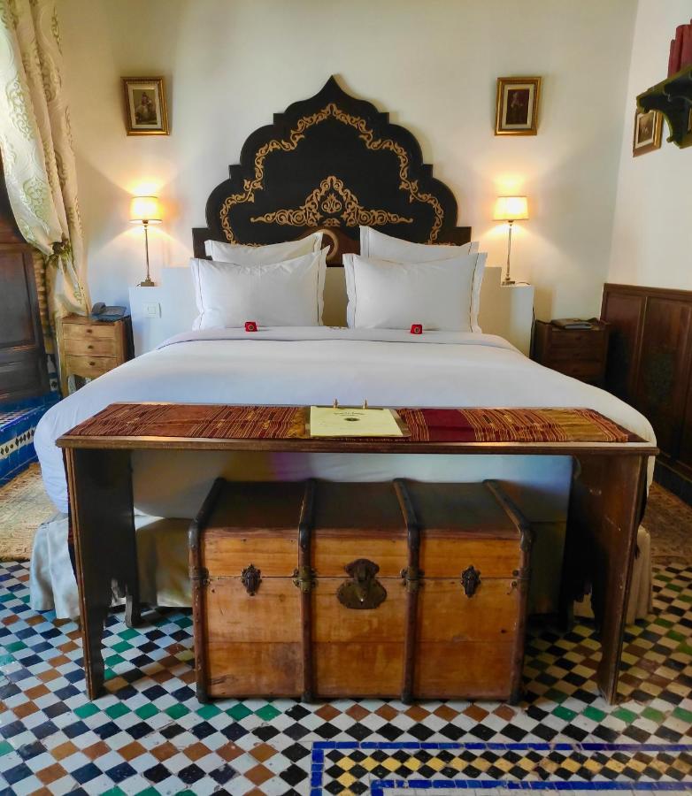 7 UNIQUE Riads in Fez You Won't Forget 19 A cozy bedroom reminiscent of riads in Fez, with a large bed featuring white linens, an ornate black and gold headboard, bedside lamps, framed art, a wooden desk, and an antique chest on a patterned tile floor.