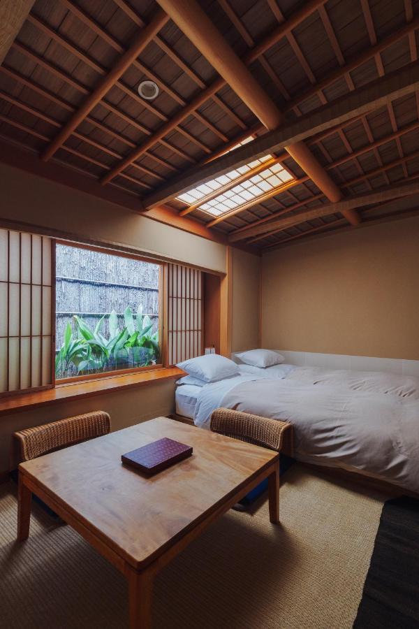 7 UNIQUE Ryokans in Gion Kyoto 8 A cozy Japanese-style room inspired by ryokans in Gion Kyoto, featuring a low wooden table, two wicker chairs, a large bed, tatami flooring, and shoji screens by the window overlooking a lush garden.