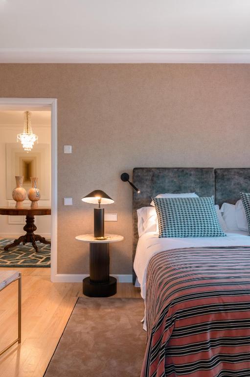 11 UNIQUE Boutique Hotels in Tenerife 43 A cozy bedroom inspired by boutique hotels in Tenerife, with a bed featuring patterned pillows and a striped blanket, a round side table with a modern lamp, and an open doorway revealing a hallway with a chandelier and decorative vases.