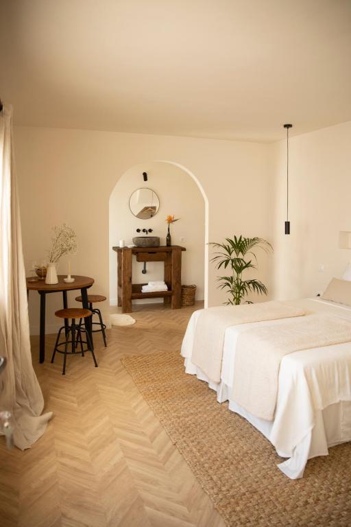 11 UNIQUE Boutique Hotels in Tenerife 15 Minimalist bedroom with light walls and a double bed with beige bedding, inspired by boutique hotels in Tenerife. A wooden table and stools sit near a window, rustic vanity with a round mirror under an arch, and potted plants add greenery.