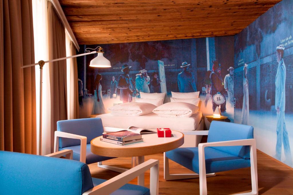 Unique Boutique Hotels in Salzburg 32 A cozy hotel room in one of the Boutique Hotels in Salzburg features a bed with white bedding, a round table with magazines, two blue chairs, a floor lamp, and a vintage-themed wall mural. Wooden ceilings and large windows fill the space with natural light.