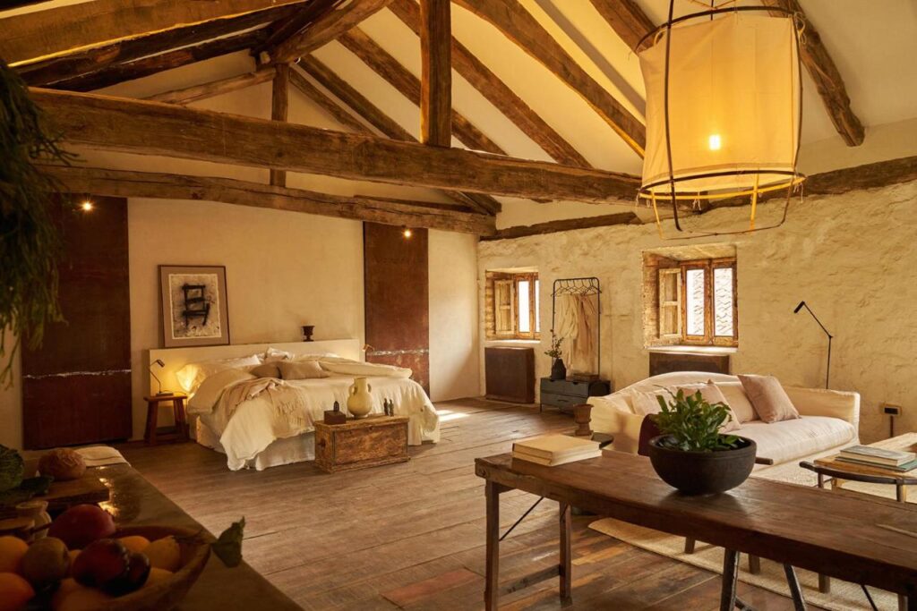 A rustic, cozy bedroom in the style of boutique hotels in Asturias, with exposed wooden beams, a large bed with light bedding, a sofa, wooden furniture, potted plants, and warm lighting from a large hanging lamp. Stone walls and wood floors complete the charm.