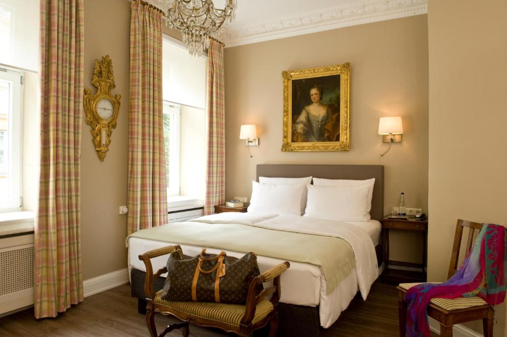 Authentic Boutique Hotels in Munich 18 A well-lit hotel room in one of the finest boutique hotels in Munich, featuring a double bed, elegant chandelier, classic decor, tall windows with plaid curtains, a gold-framed portrait above the bed, and a bag on a bench at the foot of the bed.