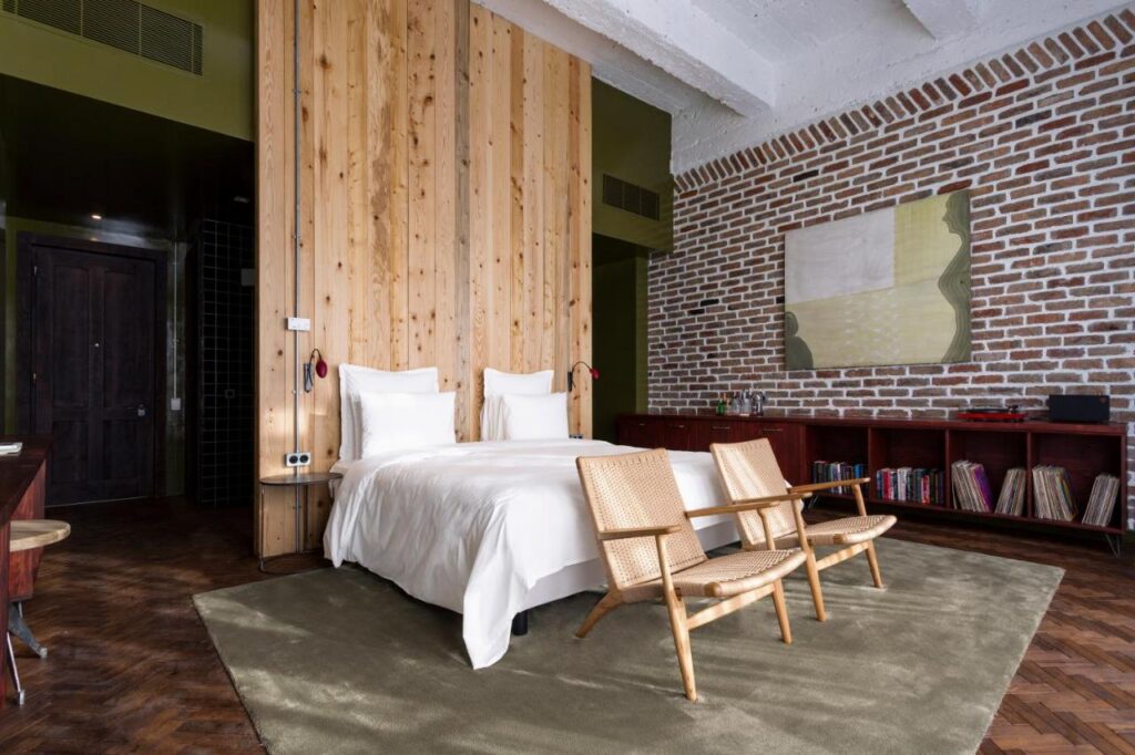 11 Unique Hotels in Georgia You Can't Miss! 4 A modern bedroom, inspired by unique hotels in Georgia, features a double bed with a wooden headboard, wicker chairs, a green rug, exposed brick walls, and a long shelf with books and decor beneath an abstract painting.