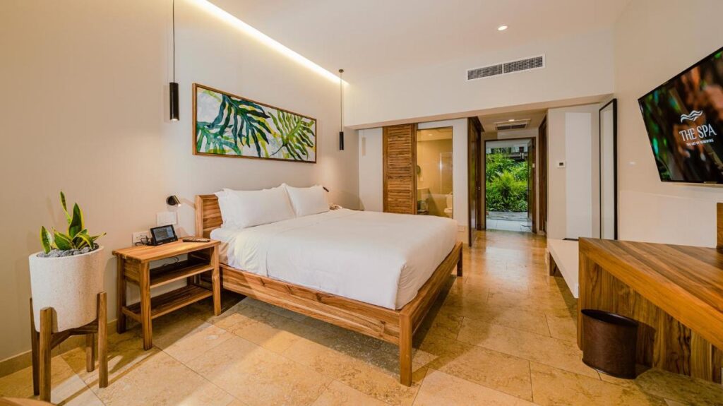 Unique Hotels in Costa Rica: Top Stays 4 A modern hotel room in one of the unique hotels in Costa Rica, featuring a large bed, wooden furniture, a potted plant, wall art with green leaves, a flat-screen TV, and serene views of greenery through a glass sliding door.