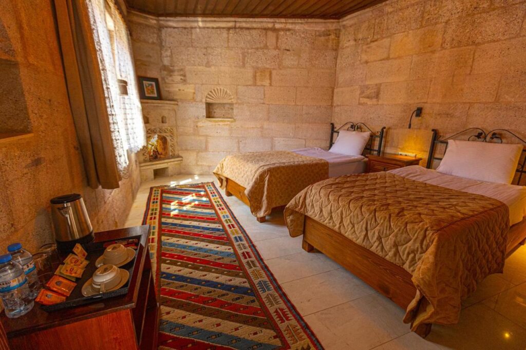 Most Authentic Cave Hotels in Cappadocia 5 A cozy stone-walled bedroom, reminiscent of cave hotels in Cappadocia, with two single beds, a colorful patterned rug, sheer-curtained window, nightstand, and a small table set with bottled water, cups, tea, and a kettle. Warm sunlight streams in.