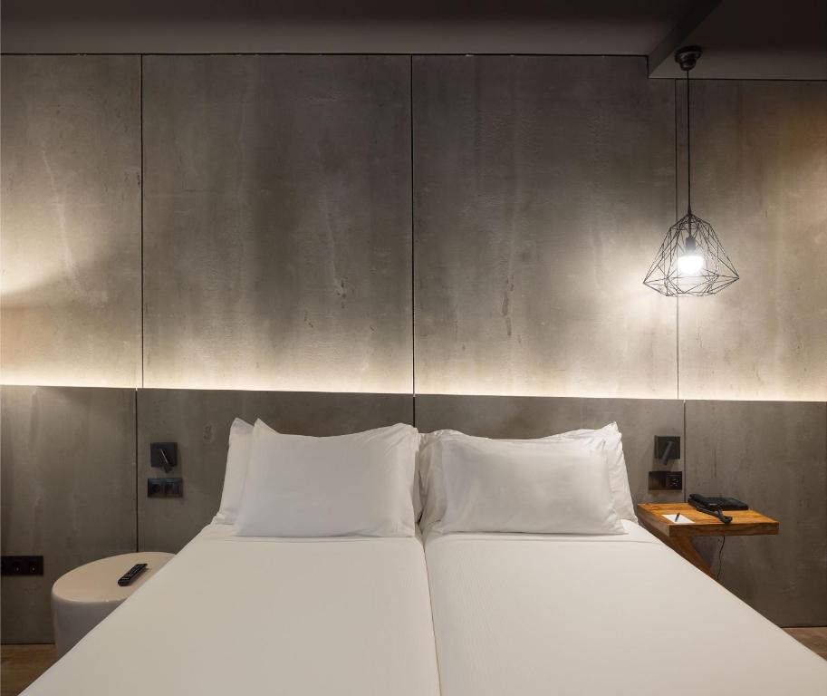 8 Unique Boutique Hotels in Bilbao Spain 6 A minimalist hotel room typical of boutique hotels in Bilbao, featuring two single beds pushed together, white bedding, a concrete accent wall, a hanging wire lamp, and cozy soft lighting for a warm atmosphere.