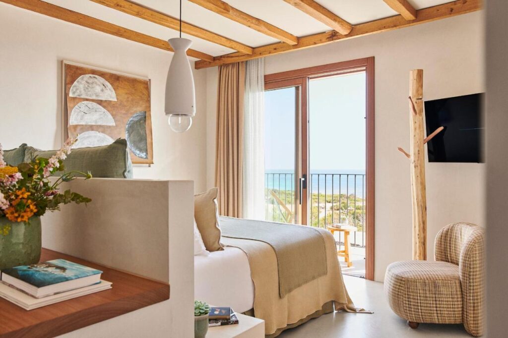 A cozy, sunlit bedroom in the style of boutique hotels in Formentera, with a double bed in neutral tones, a wooden beam ceiling, modern decor, wall-mounted TV, and sliding glass doors opening to a balcony with an ocean view.