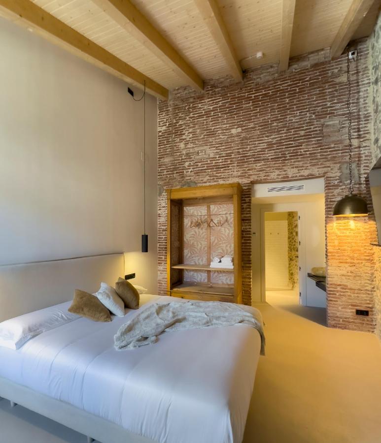 A cozy bedroom inspired by boutique hotels in Costa Brava, with exposed brick walls, wooden ceiling beams, a large bed with white linens and brown pillows, warm lighting, and an open closet space near the doorway.