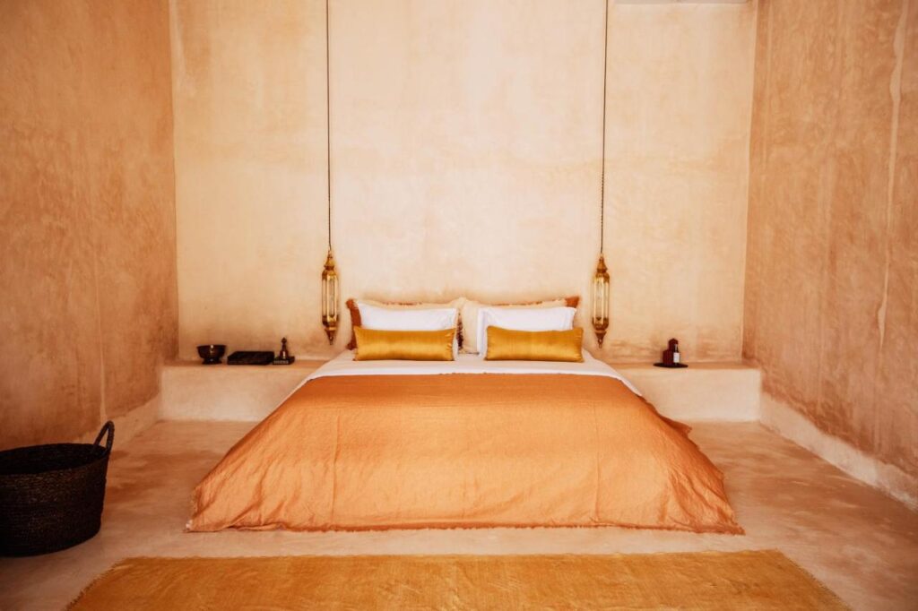A minimalist bedroom, inspired by the best family villas in Bali, features tan walls, an orange bedspread, yellow accent pillows, pendant lights, a yellow rug, simple shelf decor, and a woven basket on the floor.
