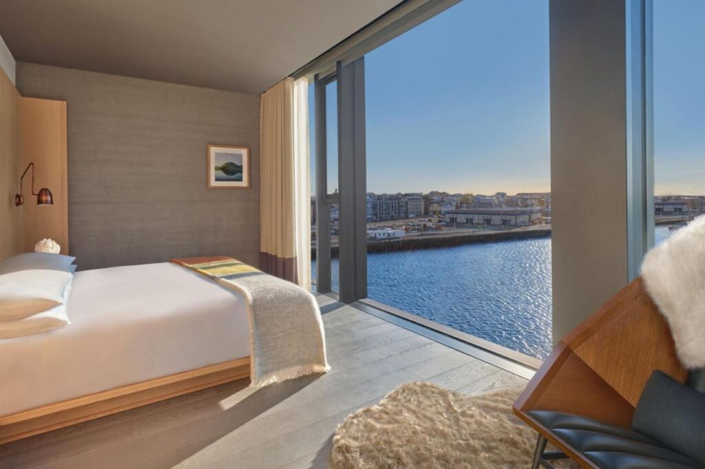 Where to Stay in Reykjavík: Top Guide 2 A modern hotel room with a large window offers a scenic view of water and city buildings—an ideal option for those seeking where to stay in Reykjavik. The room features a neatly made bed, fur-accented chair, and minimalist decor in neutral tones.