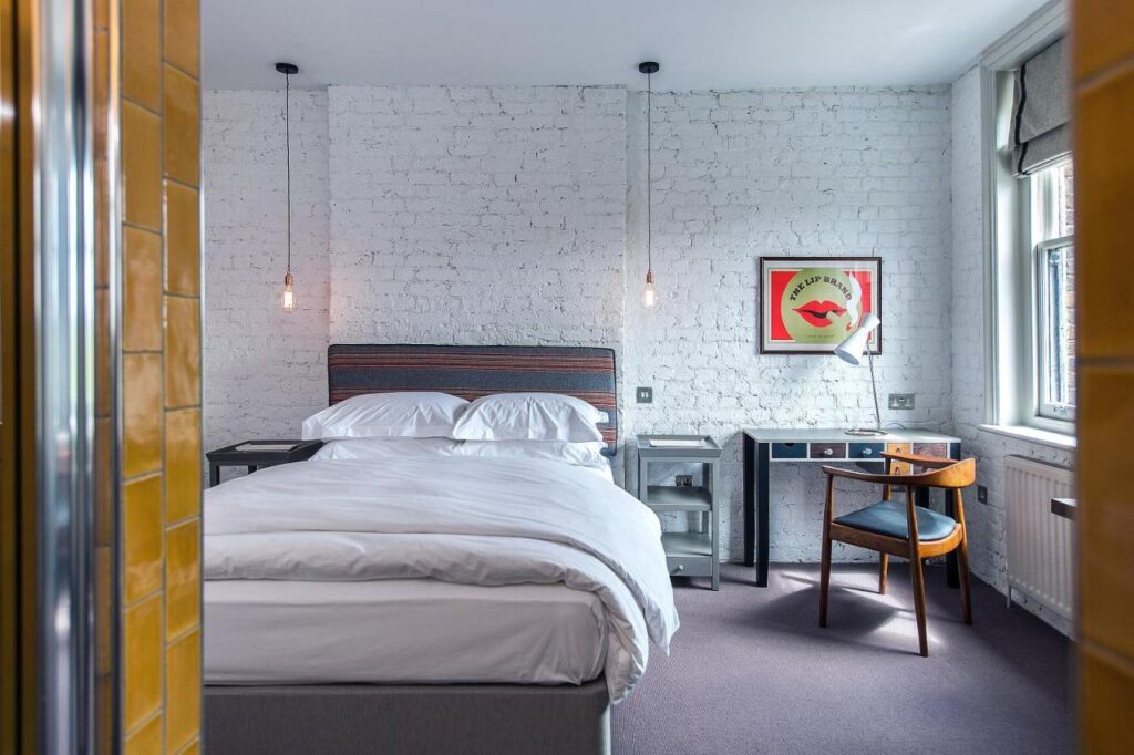15 Best Boutique Hotels in London, UK 14 A modern bedroom inspired by the best boutique hotels London offers, featuring a double bed with white bedding, exposed white brick wall, hanging lights, desk and chair, framed art, and a large window flooding the space with natural light.
