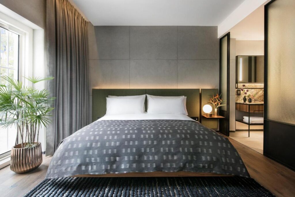 13 Most Unique Hotels in Singapore for an Incredible Holiday 19 A modern bedroom in one of the unique hotels in Singapore features a large bed with gray bedding, white pillows, a plant by a window with sheer curtains, a rug, minimalistic decor, and a partially open door leading to the bathroom.