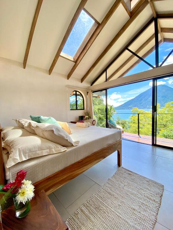 8 UNIQUE Places Where to Stay in Lake Atitlán 26 Bright, airy bedroom with wooden beams, a skylight, and large glass doors opening to a balcony with scenic lake and mountain views—an ideal choice for those seeking where to stay in Lake Atitlan. Neutral bedding and fresh flowers complete the space.