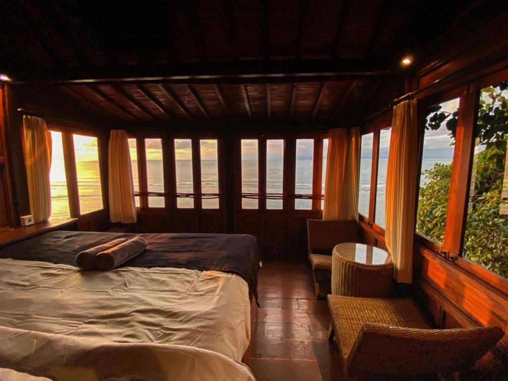 A cozy wooden bedroom in one of the best family villas in Bali features large windows with panoramic ocean sunset views. A double bed, two wicker chairs, and a small table are bathed in warm, golden light.