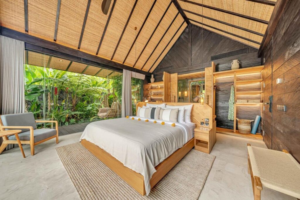 14 UNIQUE Boutique Hotels in Canggu, Bali 7 A cozy, modern bedroom inspired by boutique hotels in Canggu features a wooden vaulted ceiling, large bed with white linens, tropical decor, armchairs, and glass doors opening to lush greenery. Warm natural light fills the spacious room.