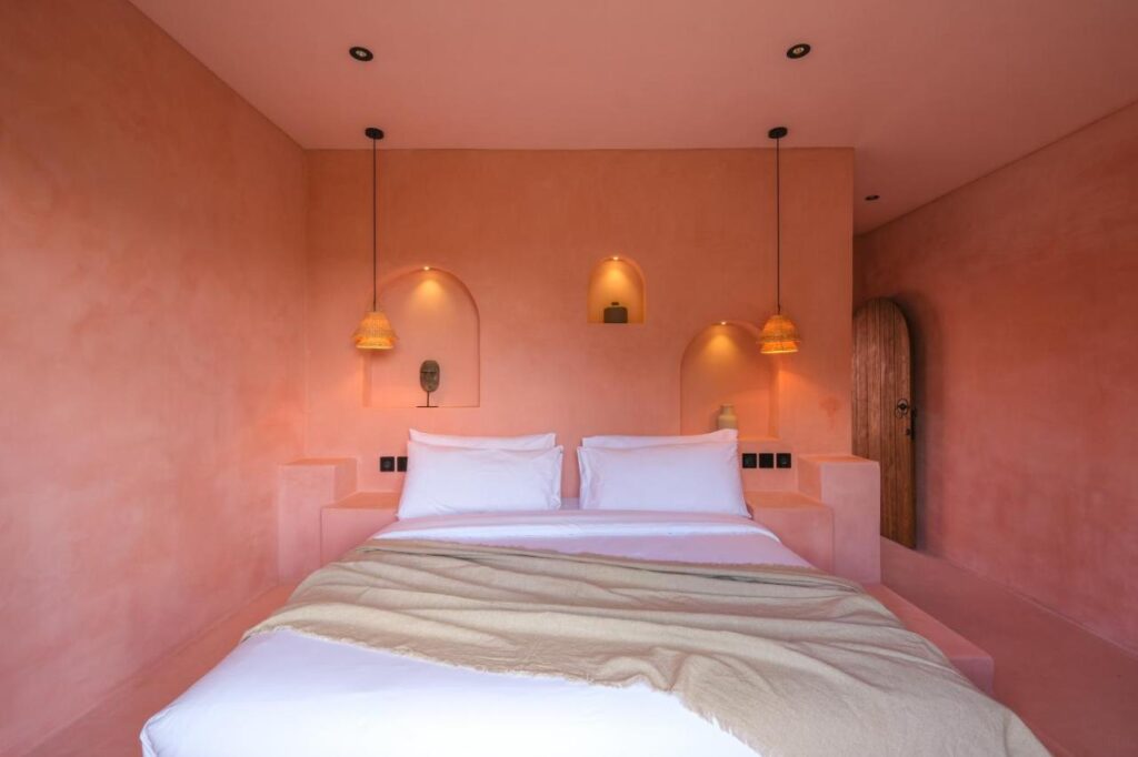 A cozy bedroom in one of the best family villas in Bali, featuring pink walls, a double bed with white bedding, two hanging wicker lamps, and three alcoves above the bed displaying decorative objects. A wooden arched door is on the right.