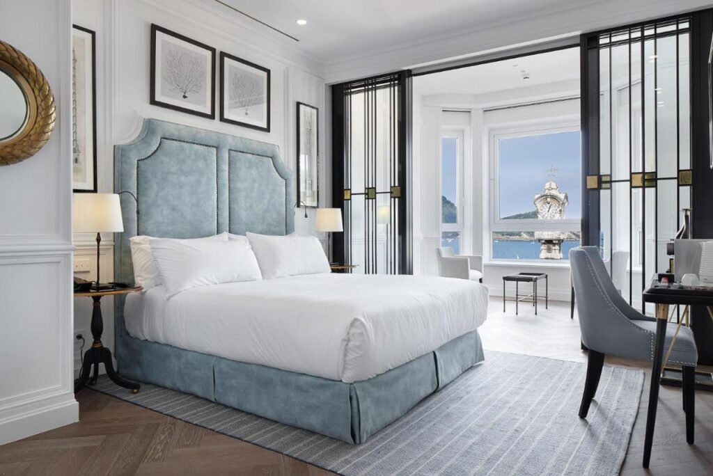 Best Boutique Hotels in San Sebastian 17 A bright, elegant hotel room with a large bed, soft blue headboard, two bedside lamps, desk, and armchair. Modern and sophisticated decor complements the scenic view—perfect for those seeking boutique hotels in San Sebastian.
