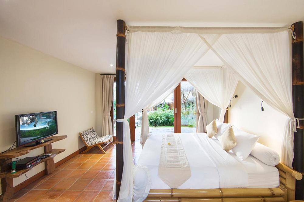 Bright, airy bedroom with a bamboo canopy bed draped in white sheer fabric, white bedding, a TV on a wooden stand, patterned chair, terracotta tile flooring, and glass doors leading to a green garden—perfect for the best family villas in Bali.