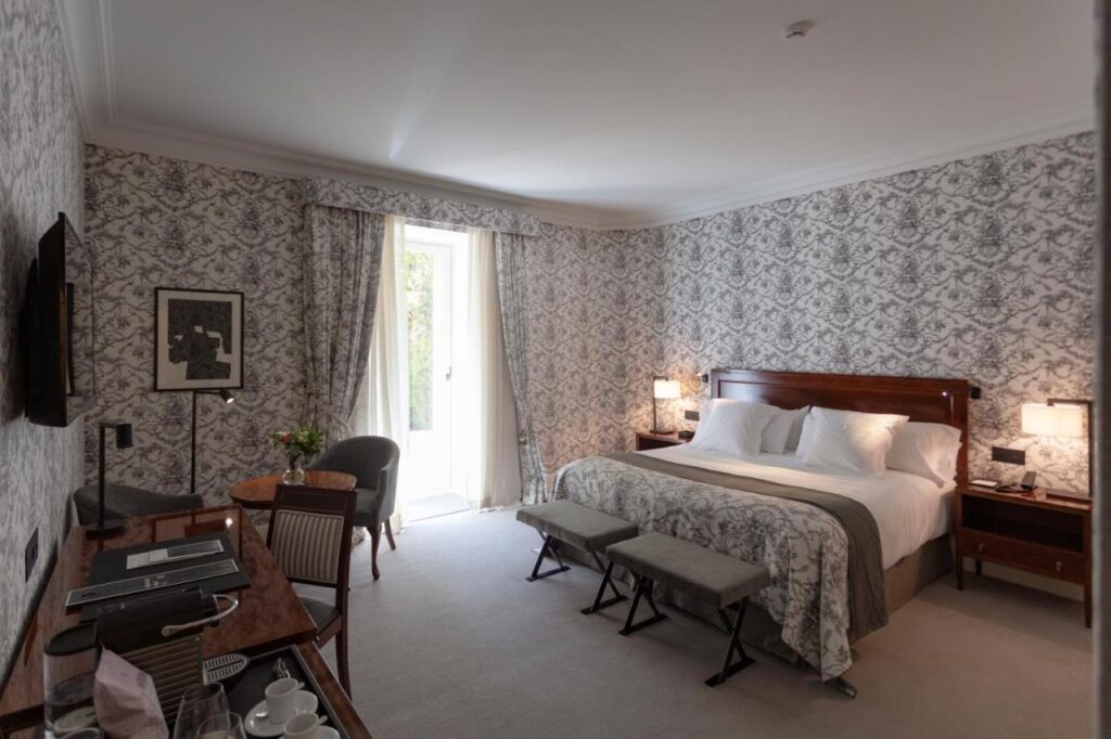 Best Boutique Hotels in San Sebastian 12 A well-lit boutique hotel room in San Sebastian features a large bed with white linens, two bedside tables with lamps, padded benches at the foot, a desk, chairs, tea set, patterned wallpaper, and a window with curtains.