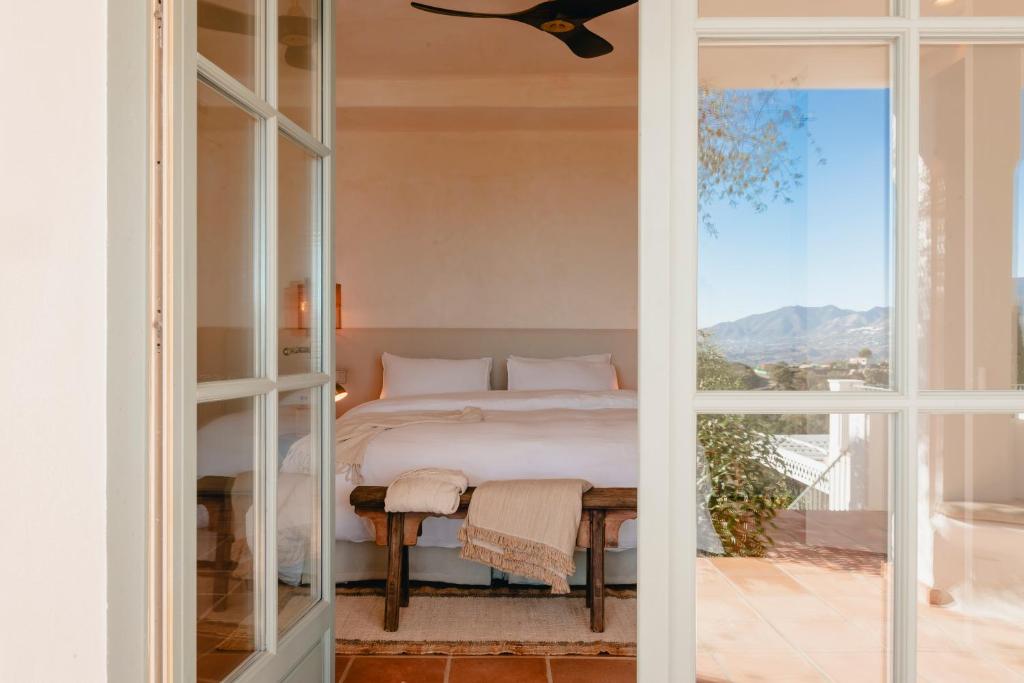 Adults-Only Hotels in Costa del Sol 11 A cozy bedroom at one of the adult only hotels in Costa del Sol, featuring a large bed with white linens, a wooden bench, and a light throw blanket. Sunlight streams in through glass doors, revealing mountain views in the background.