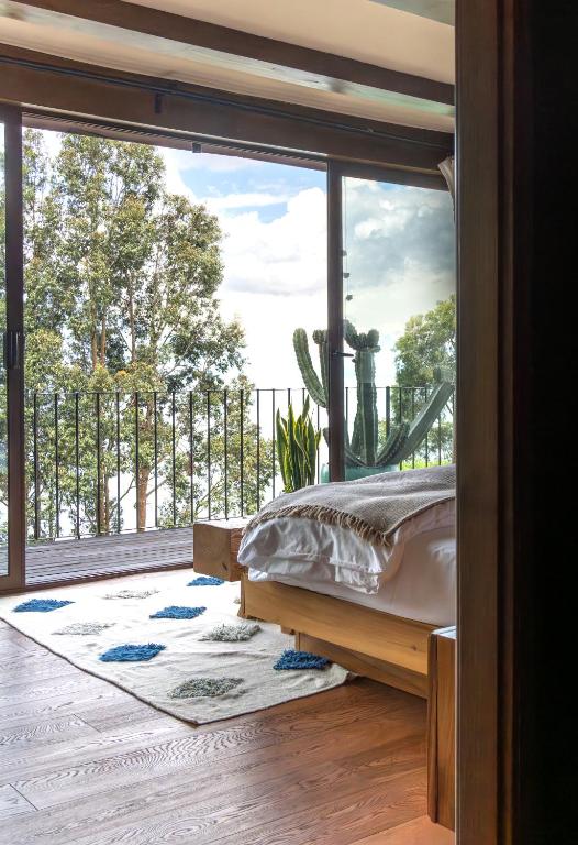 8 UNIQUE Places Where to Stay in Lake Atitlán 15 A cozy bedroom with a wooden floor and bed, a white rug with blue patterns, and large glass doors opening to a balcony with trees and sky visible outside—perfect for those seeking where to stay in Lake Atitlan.