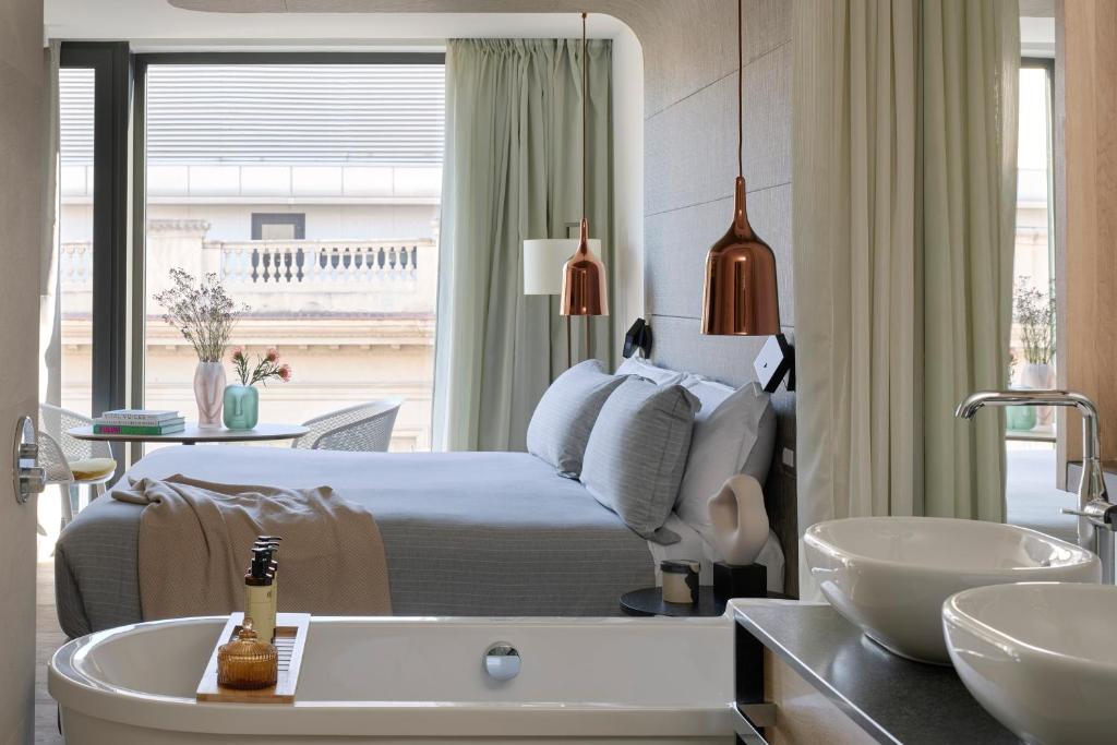 Top Barcelona Hotels with Rooftop Pool 27 A modern hotel room with a double bed, light gray bedding, copper pendant lights, and a bathtub with twin sinks. Large windows with sheer curtains overlook a balcony—perfect for those seeking Barcelona hotels with rooftop pool access.